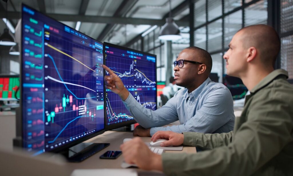 Zwei Personen analysieren Börsencharts mit Kerzenmustern und Kurslinien auf mehreren Monitoren.