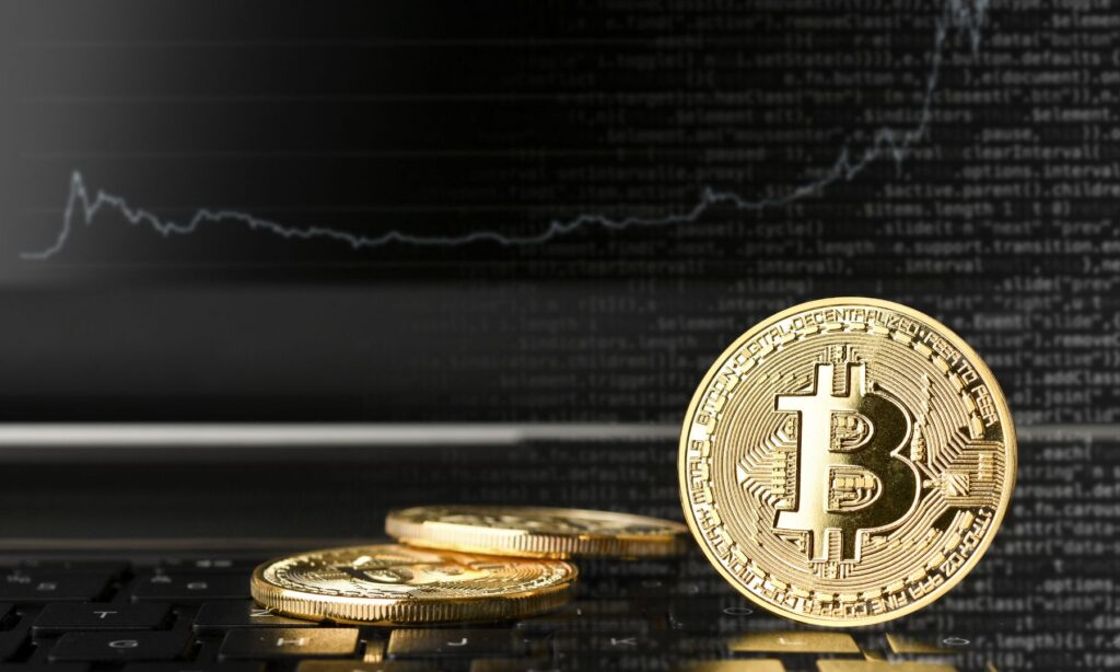 Drei goldene Bitcoin-Münzen auf einer Tastatur vor einem Bildschirm mit Programmcode und Kurslinie.